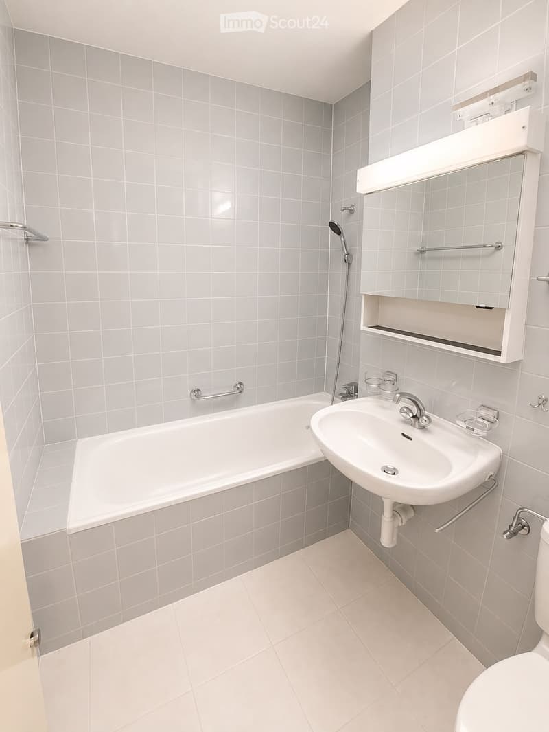Modern bathroom with white tiled walls and floor, featuring a bathtub, sink, mirror, and toilet.