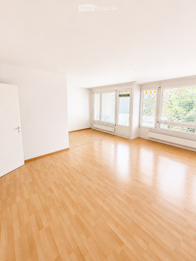 Empty room with light wooden flooring, white walls, large windows with blinds, and a door leading outside.
