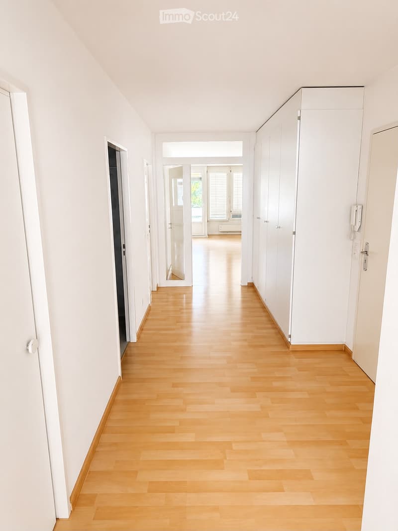 Bright, empty hallway with light wooden flooring and white walls, featuring several doors and a view into another room at the end.