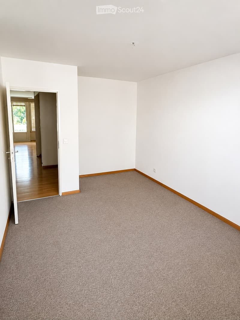 Empty room with light-colored walls, carpeted floor, and an open doorway leading to another room with wooden flooring.