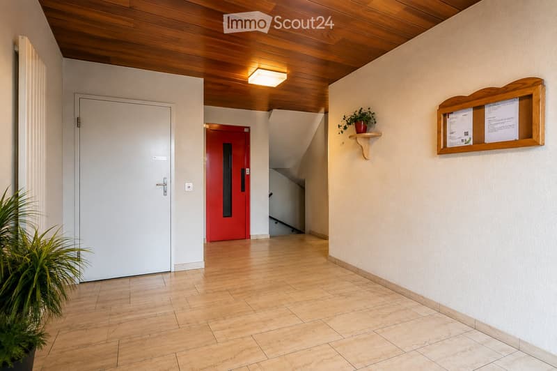 Clean, well-lit hallway with wooden flooring, white walls, and a wooden ceiling. Features include a white door, a red door leading to a staircase, a small plant on a shelf, and a bulletin board with papers.