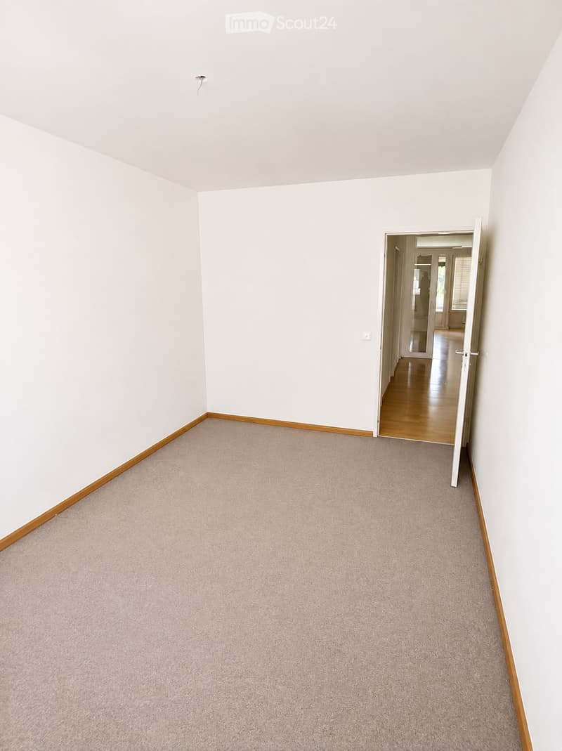 Empty room with light-colored walls, carpeted floor, and a wooden baseboard. An open doorway leads to a hallway with wooden flooring.
