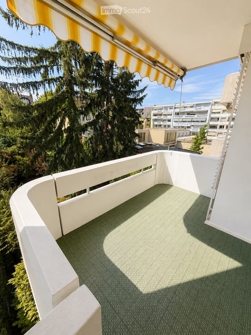 Balcony with green tiled floor, white railing, and yellow and white striped awning, overlooking a tree-lined area with buildings in the background.
