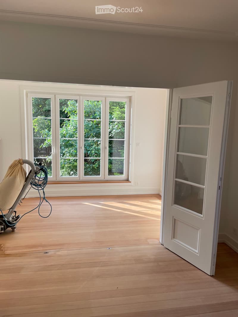 Empty room with wooden flooring, large windows with white frames, and an open door leading to another space. Sunlight streams in, illuminating the space.