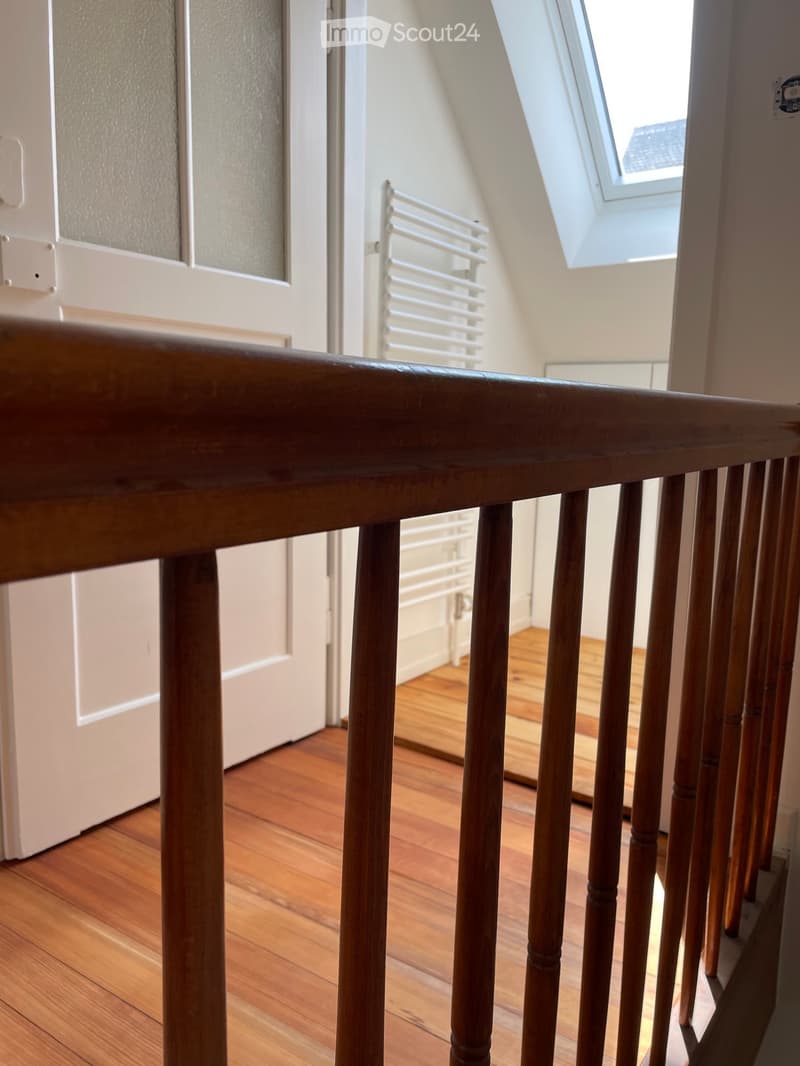 View from a staircase looking down a hallway with wooden flooring and white walls. A door is visible on the left, and a radiator is mounted on the wall in the background.