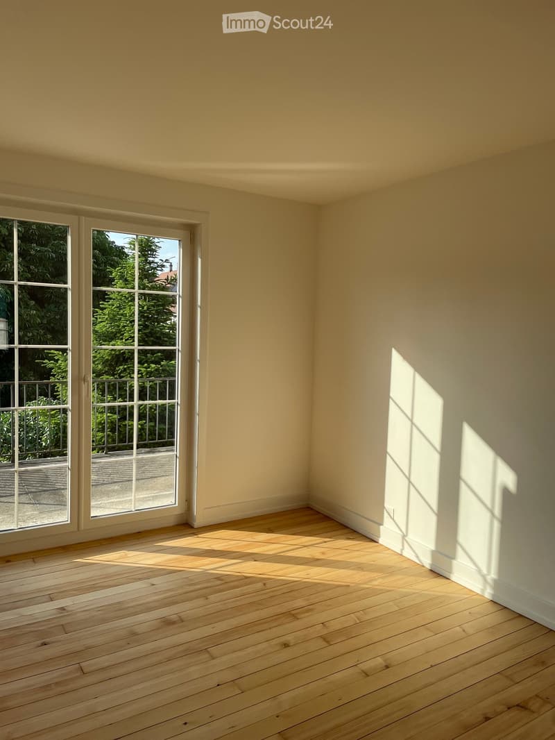 Empty room with light-colored walls and wooden flooring, featuring large windows with a view of greenery outside.