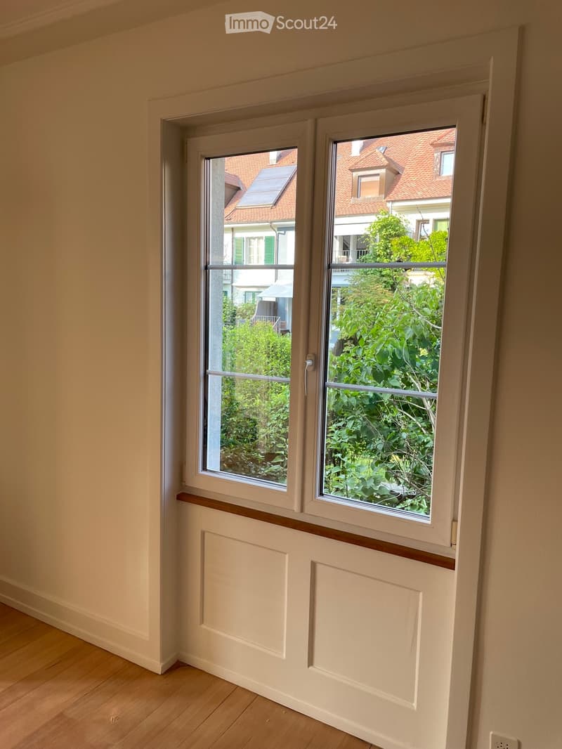 Empty room with light-colored walls, wooden flooring, and two large windows showing a view of neighboring houses and greenery.