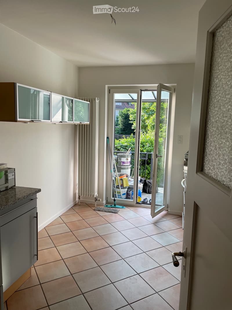 A clean, tiled room with a cabinet, radiator, and a mop leaning against the wall. An open sliding door leads to a balcony with greenery visible outside.