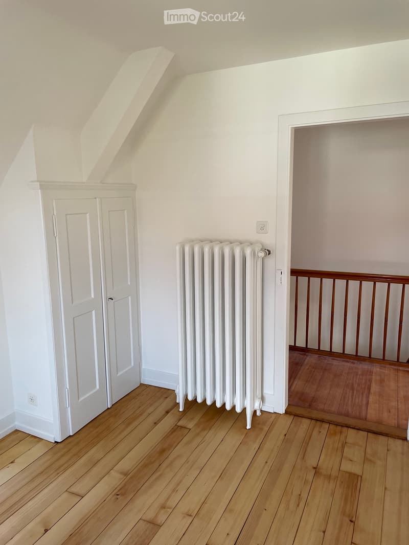 Empty room with wooden flooring, white walls, a radiator, and a white closet door. A wooden railing is visible in the adjacent area.