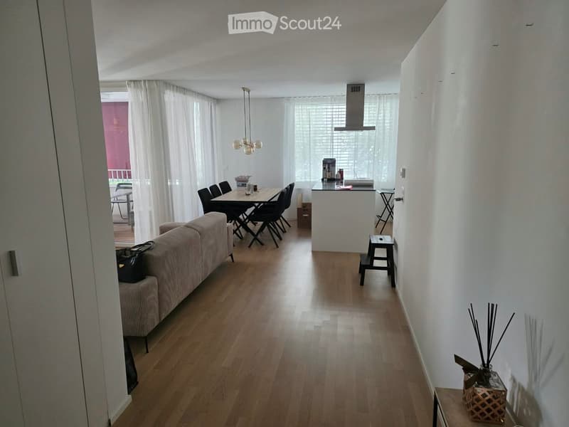Open-plan living and dining area with light wooden flooring, white walls, and large windows with white curtains. Features a sofa, dining table with chairs, a small kitchen counter, and decorative items.