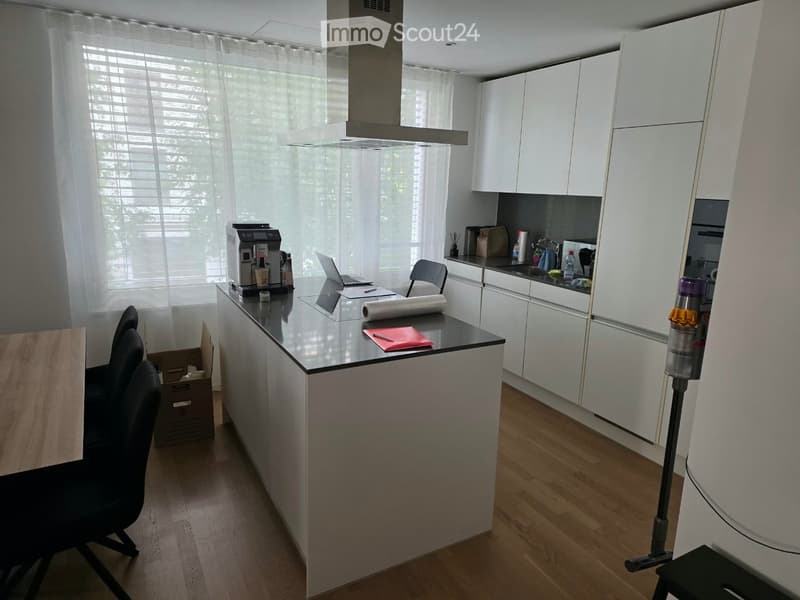 Modern kitchen with white cabinets, a central island, and a coffee machine on the counter. There is a laptop, a chair, and a small table visible.