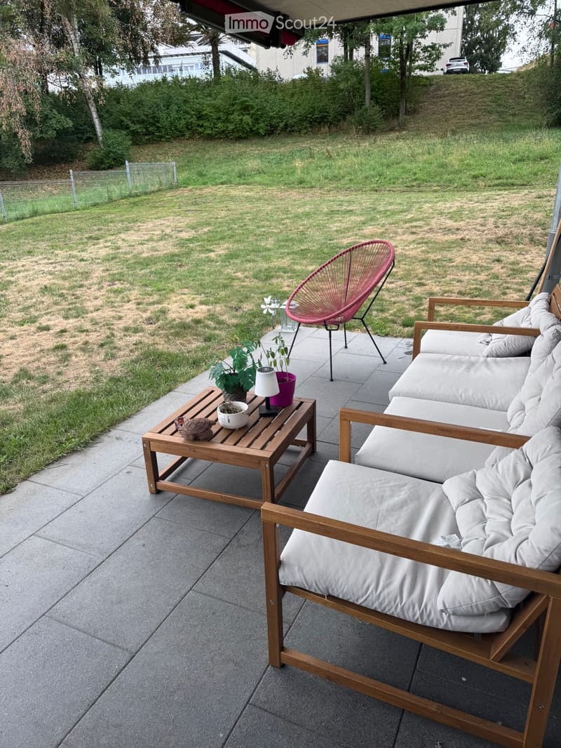 Outdoor patio with wooden furniture, including a sofa, chairs, a table, and a lamp, set on a concrete surface with a grassy area in the background.