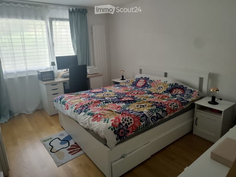 A well-lit bedroom with a bed, desk, and nightstand. The bed has a colorful floral duvet, and the room features wooden flooring and white walls.