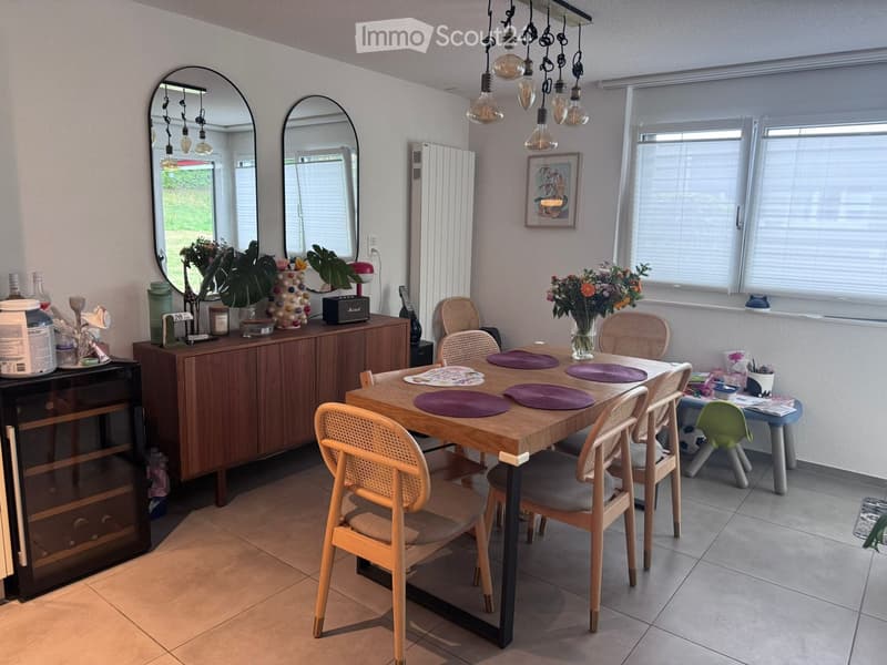 Dining room with a wooden dining table surrounded by chairs, a cabinet with decorative items, mirrors, and a vase with flowers on the windowsill.