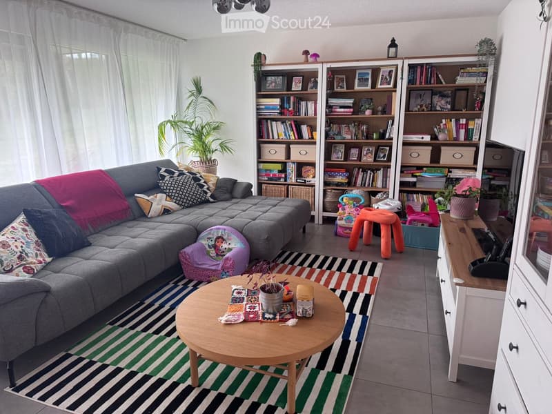 Living room with grey sectional sofa, colorful cushions, coffee table, bookshelf, and small orange stool.