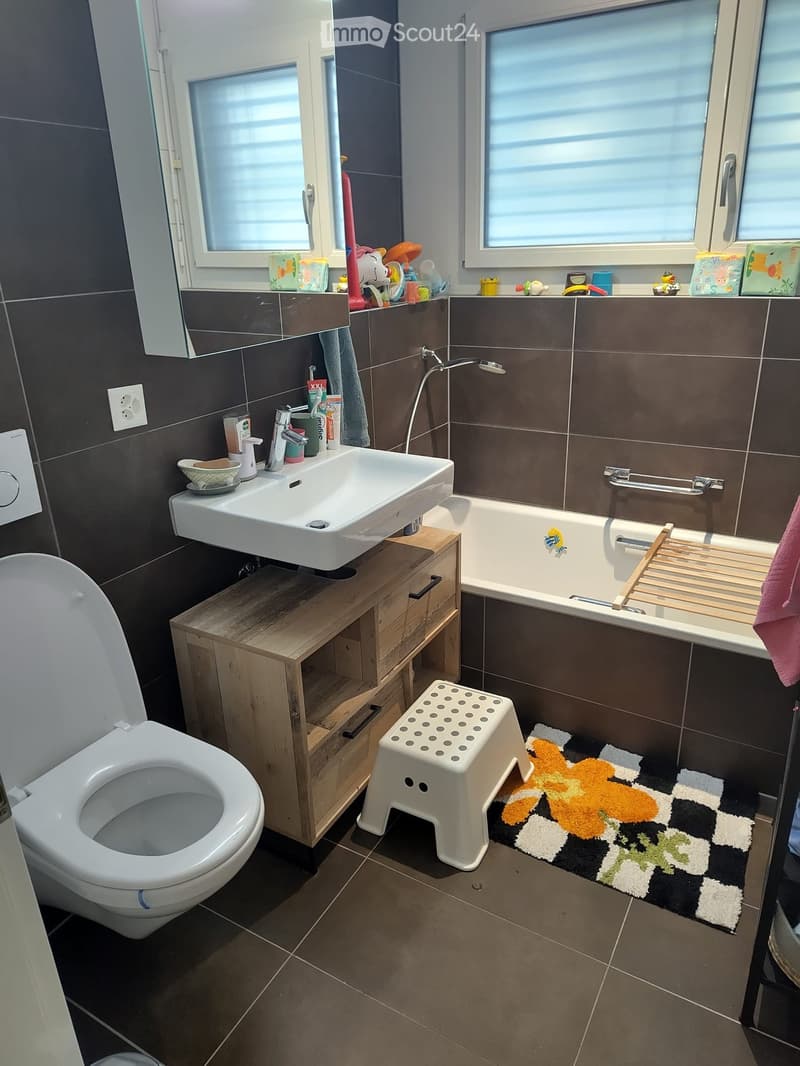Modern bathroom with dark tiled walls, a white sink, a toilet, a bathtub, a wooden shelf, a small white stool, and a colorful bath mat.