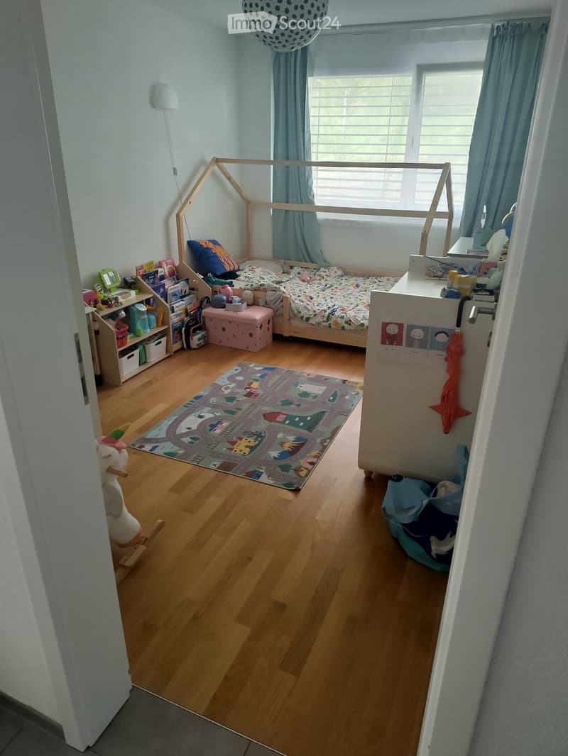 Child's bedroom with a wooden bed frame, colorful bedding, a shelf with books and toys, a pink stool, a play rug, and a cat bed.