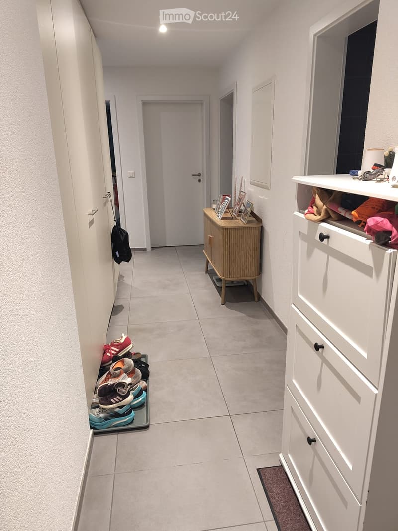 Clean, modern hallway with tiled flooring, white walls, and several doors. Features include a small wooden side table with framed photos, a white storage cabinet with drawers, a shoe rack with multiple pairs of shoes, and a coat hook with a black jacket h