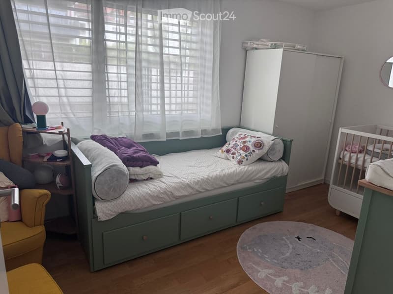 Cozy bedroom with a single bed, nightstand, small chair, and a baby cradle. The room features a wooden floor, a round rug, and a window with sheer curtains.