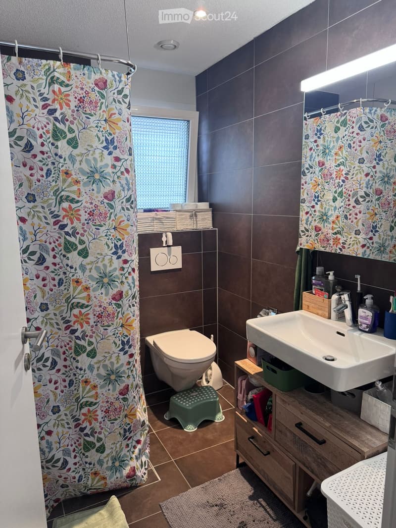 Modern bathroom with dark brown tiles, a white toilet, a sink with a wooden cabinet, a shower with floral curtains, and a small green stool.