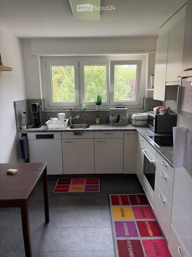 Modern kitchen with white cabinets, granite countertops, and stainless steel appliances. Features a sink, oven, refrigerator, microwave, coffee maker, mixer, and toaster. A small table with a box on it is visible in the foreground.