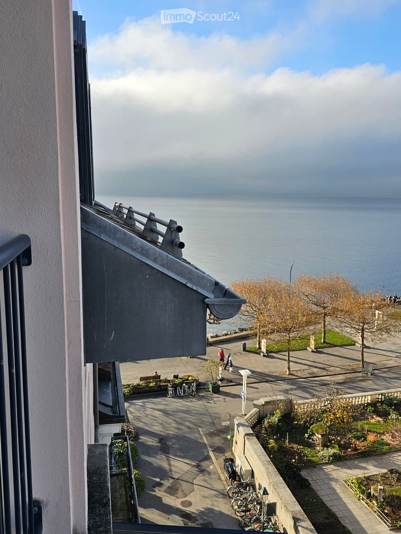 Balcony overlooking a waterfront area with a paved walkway, trees, and a few people walking.