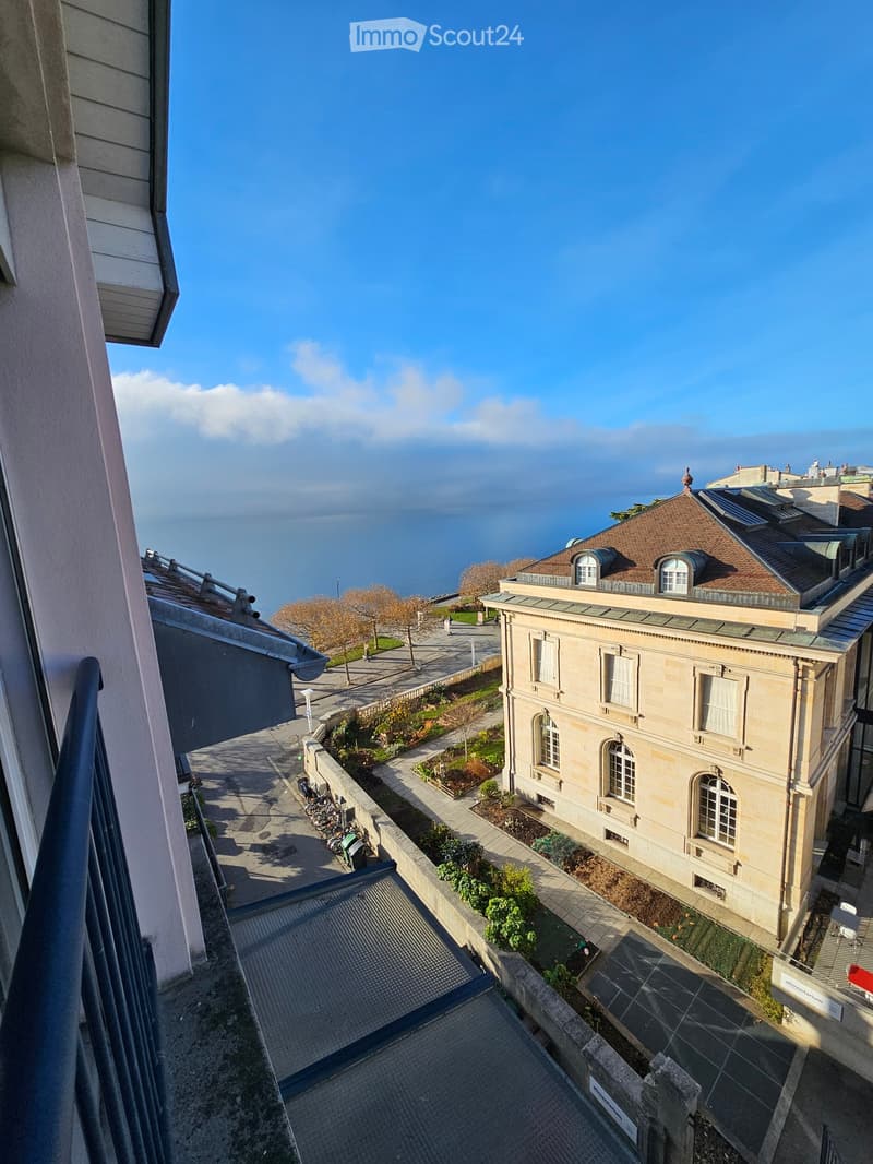 Balcony overlooking a waterfront with a historic building, garden area, and a clear view of the lake and distant shoreline.