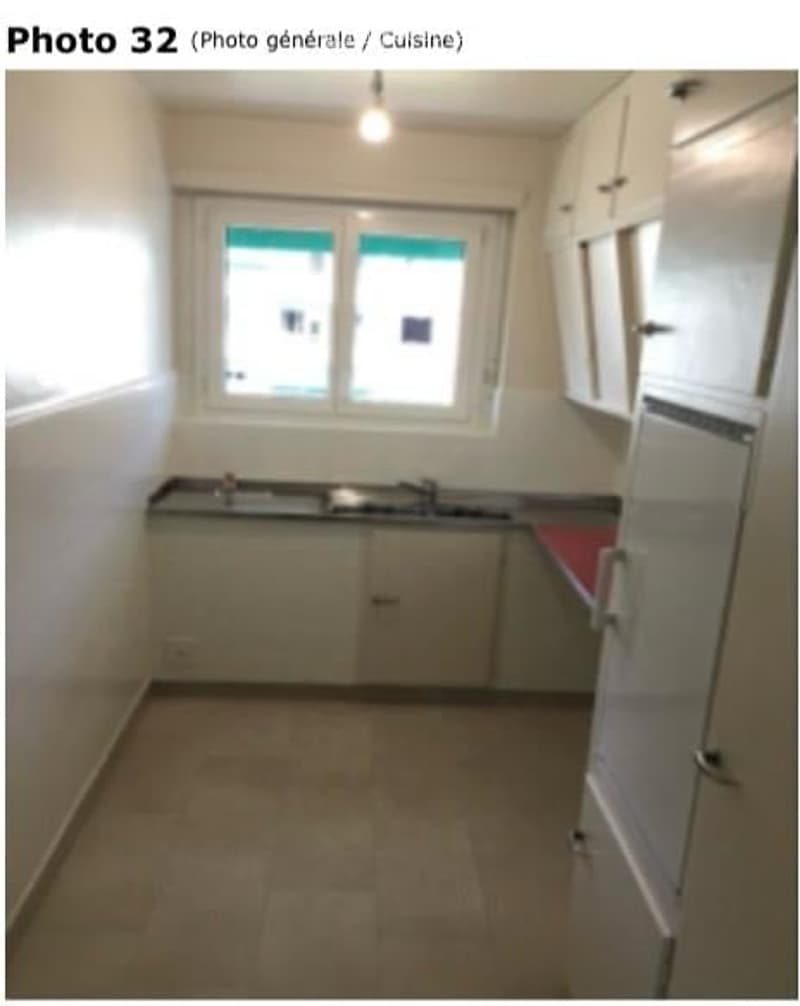 A small, unfurnished kitchen with white walls, a window, a sink, and cabinets. The floor is tiled, and there is a refrigerator on the right side.