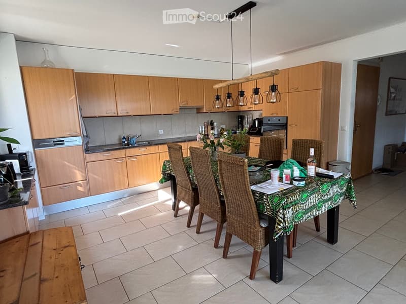 Bright kitchen with light wooden cabinets, a central dining table with wicker chairs, and various appliances including a sink, oven, and dishwasher.