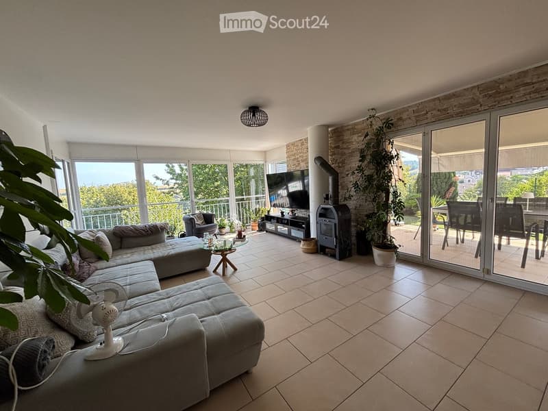 Bright living room with large windows and sliding glass doors leading to a balcony. Features a grey sectional sofa, coffee table, fan, TV, and additional seating.