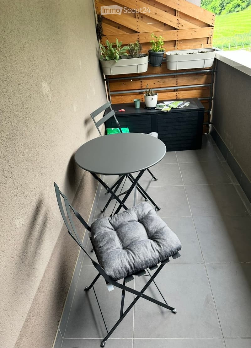 Balcony with grey tiled floor, round folding table, folding chair with grey cushion, storage box, and several potted plants on a wooden shelf.