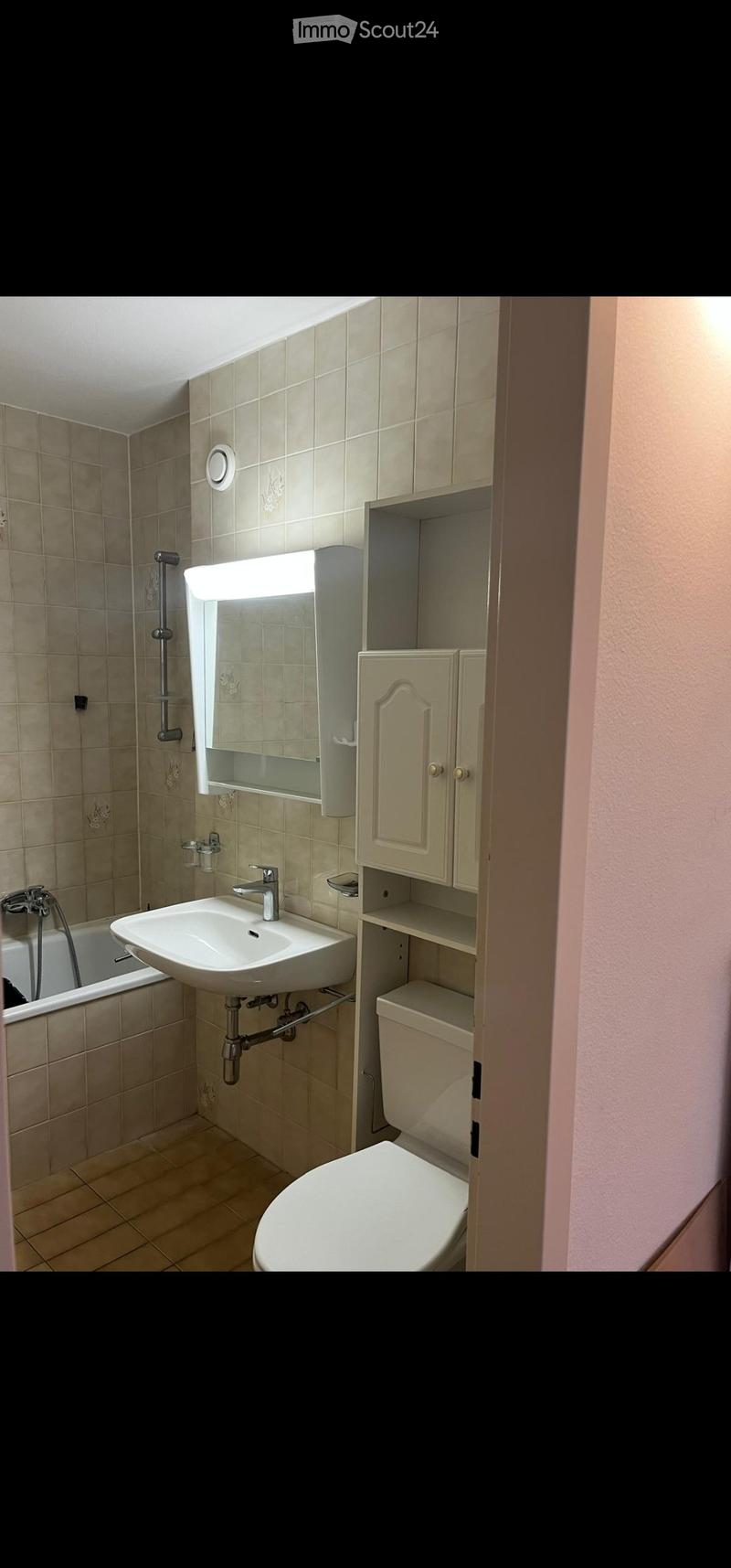 Bathroom with beige tiles, a white sink, a toilet, a bathtub, a mirror with built-in lighting, and a cabinet with shelves.