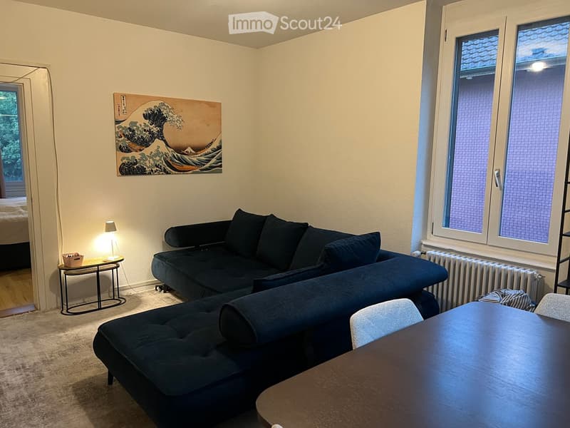 A modern living room with a dark blue sectional sofa, a small side table with a lamp, a dining table with chairs, and a large mirror on the wall.