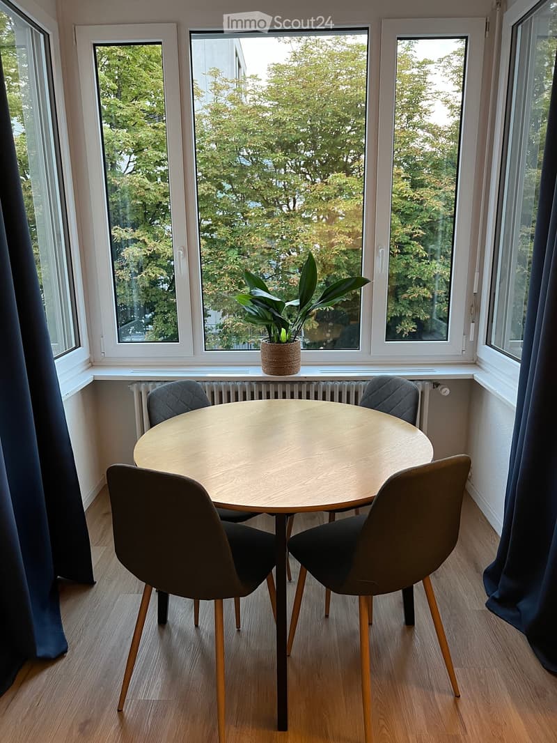 A small, well-lit room with wooden flooring, large windows, and a round wooden table surrounded by four modern chairs. A potted plant sits on the windowsill.