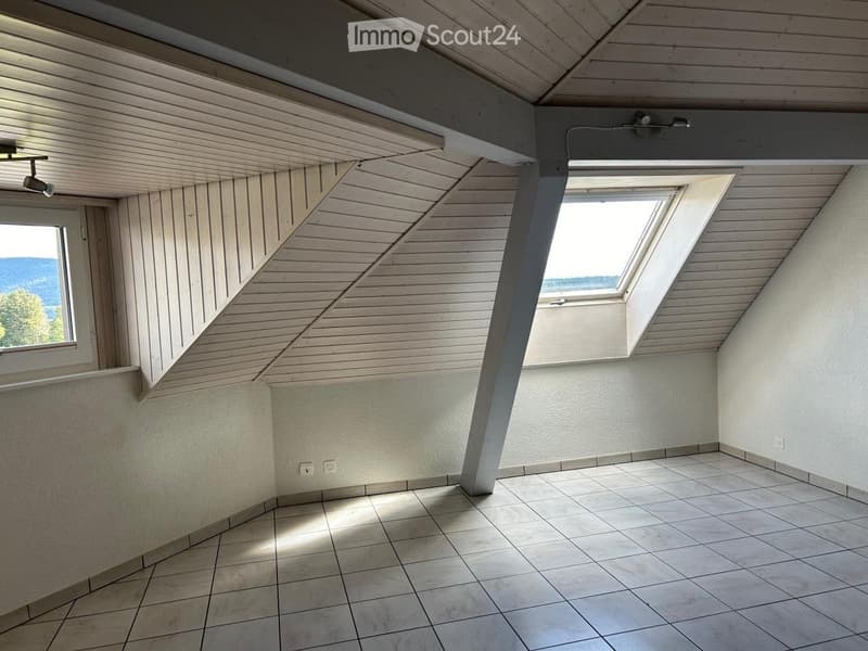 Empty attic room with wooden slatted ceiling, tiled floor, and skylight windows, featuring exposed wooden beams and natural light.