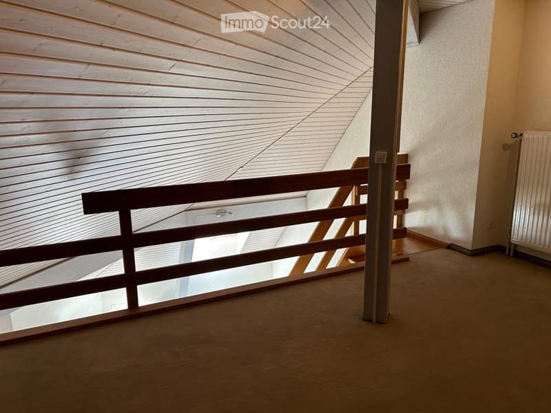 Empty room with wooden flooring, white walls, and a wooden railing leading to a raised area with a slanted wooden ceiling.