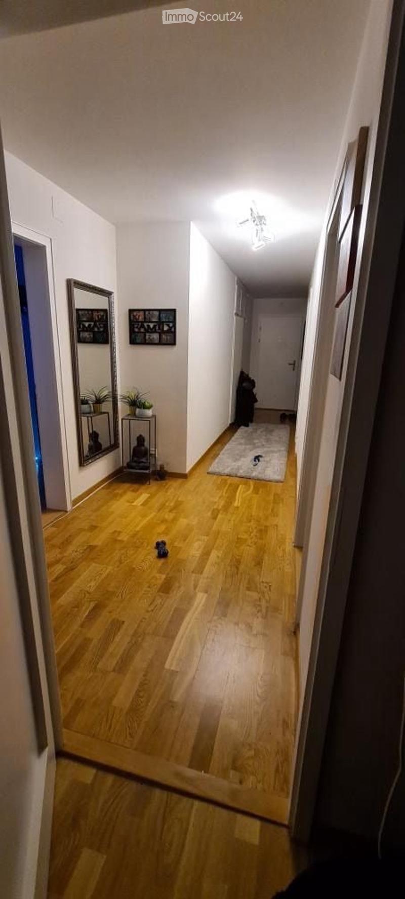 A long, narrow hallway with wooden flooring, white walls, and a ceiling light fixture. There is a small rug and a few decorative items visible along the walls.