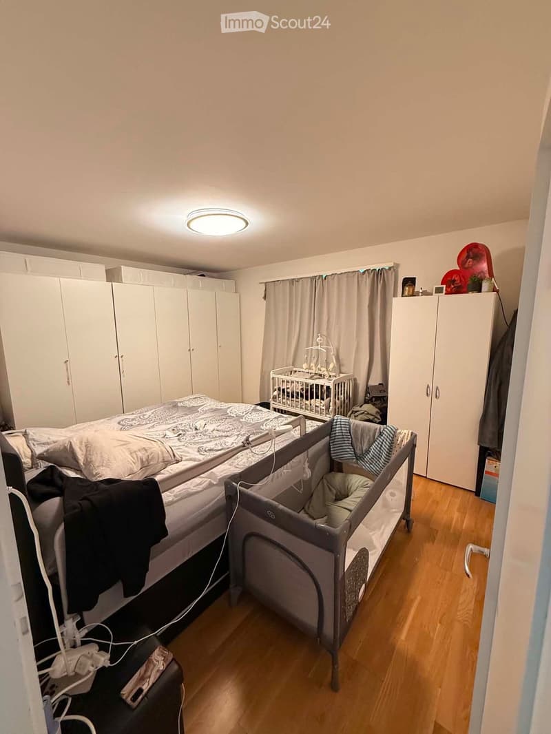 A well-lit bedroom with a double bed, a baby crib, a playpen, and built-in wardrobes. The room features wooden flooring and a ceiling light.