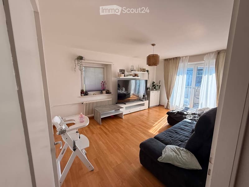 A modern living room with wooden flooring, a dark blue sofa, a coffee table, a baby chair, a television, and a large window with curtains leading to a balcony.