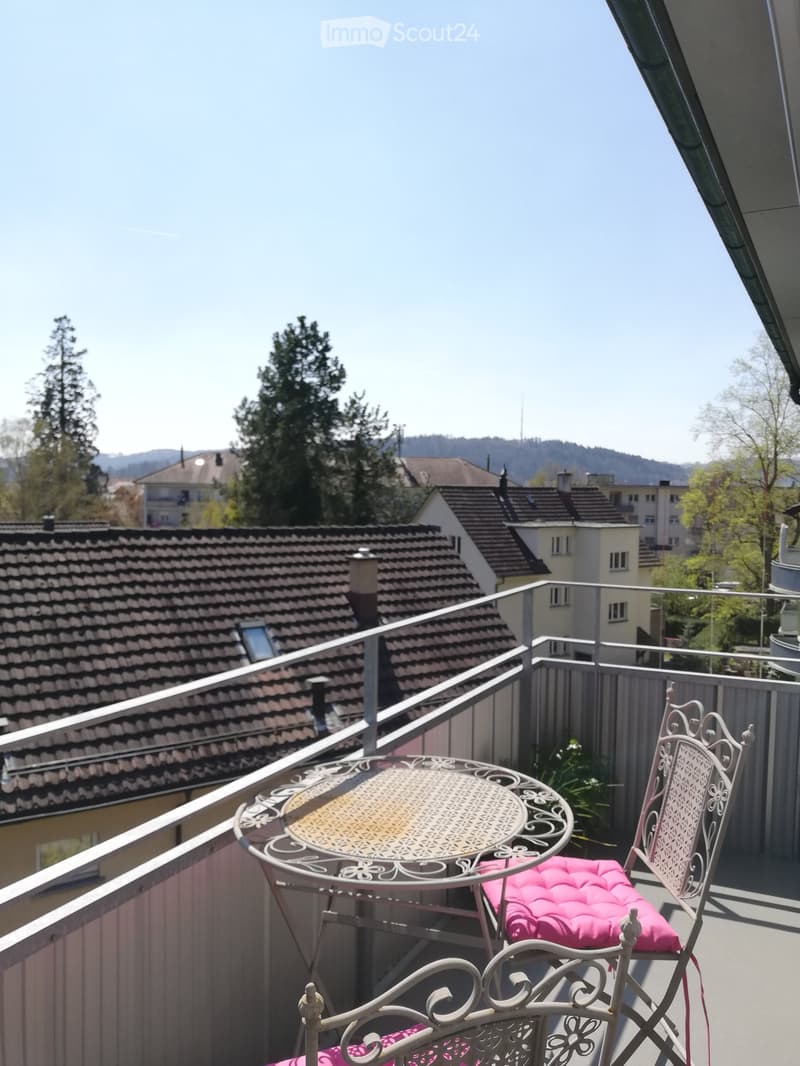 Balcony with a round metal table and two matching chairs with pink cushions, overlooking a residential area with tiled roofs and trees.