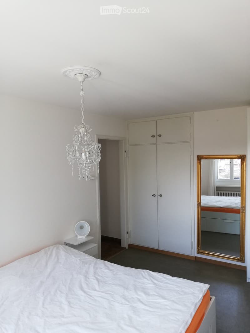 A modern bedroom with a white bed, a large wardrobe, a chandelier, a mirror, and a small table with a lamp.