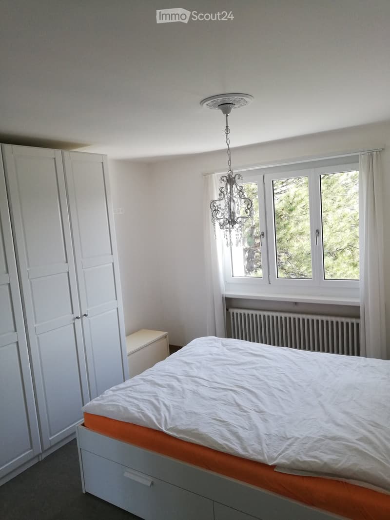 Bright bedroom with a white bed, white wardrobe, chandelier, and large windows with white curtains.