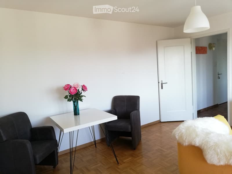 A modern living room with a white table, two chairs, a sofa, and a vase with pink flowers on the table. The room has wooden flooring and white walls.
