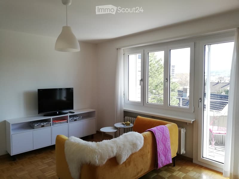 Living room with a yellow sofa, a small round table, a television on a white cabinet, and a hanging lamp. The room has large windows with a view of the city and a balcony accessible through a sliding glass door.