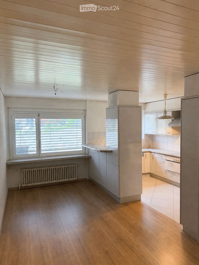 Modern kitchen with wooden flooring, white cabinets, a refrigerator, and a window with blinds. The space includes a radiator and a light fixture.