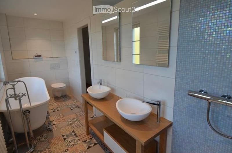 Modern bathroom with a white bathtub, toilet, two sinks on a wooden vanity, and large mirrors above the sinks. The walls are tiled in white and blue, and the floor has a patterned tile design.