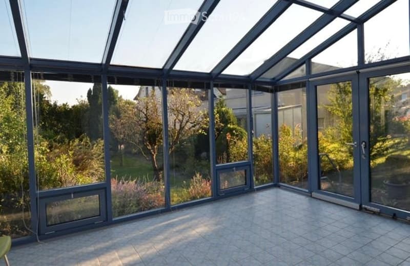 Empty sunroom with large glass walls and roof, tiled floor, and views of a garden with trees and plants.