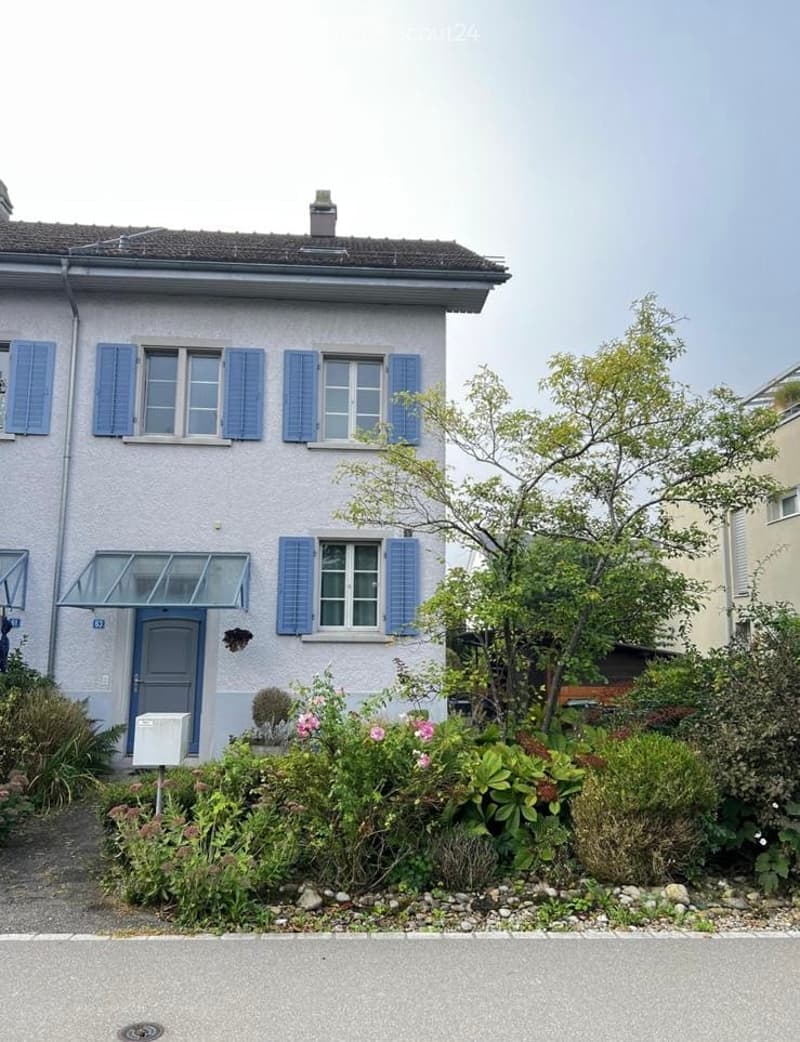 Two-story house with light grey facade, blue shutters, and a small garden with flowers and shrubs.