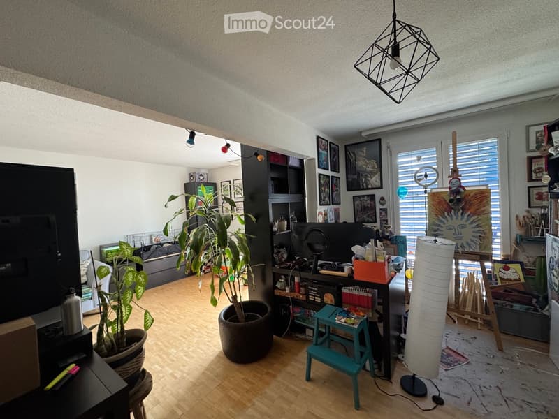 The room features a wooden floor, large windows with blinds, a hanging geometric lamp, a sofa, a TV screen, a shelf with various items, a tall plant in a pot, a small teal stool, and a tall white lamp.