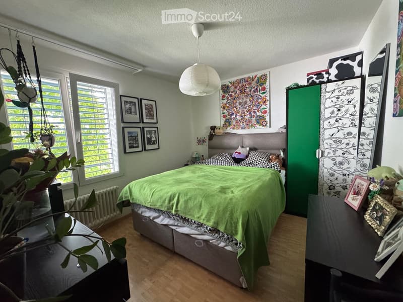 Cozy bedroom with a green duvet, wooden floor, large windows, nightstand, and decorative artwork.